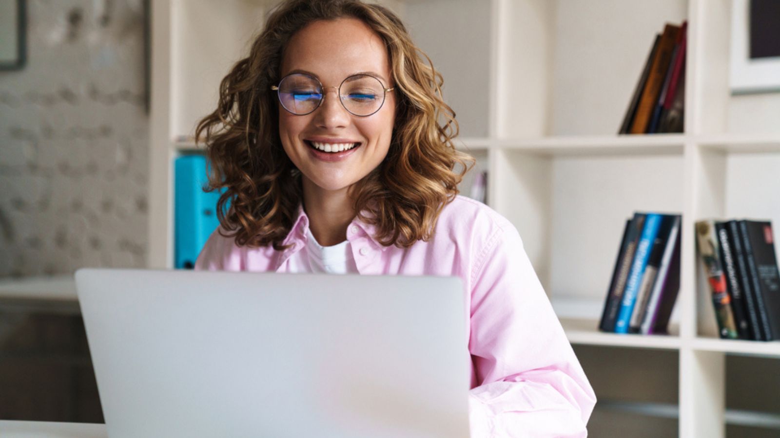 Photo of joyful blonde woman wearing eyeglasses smiling and using laptop while sitting in cafe