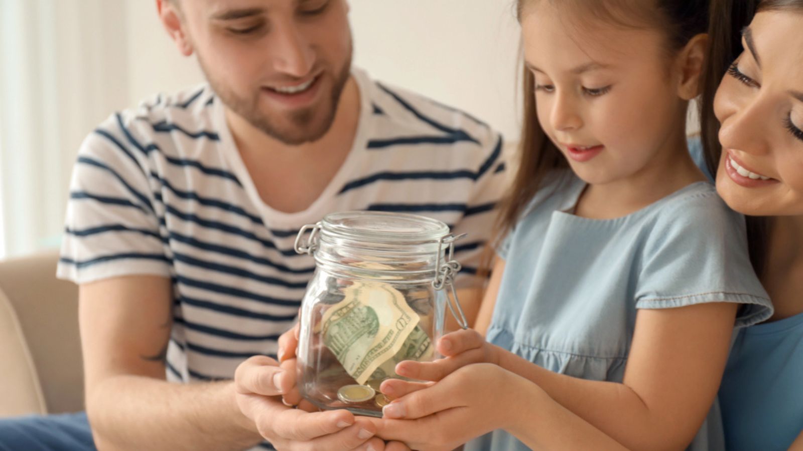Little girl with her parents holding glass jar with dollars indoors. Money savings concept