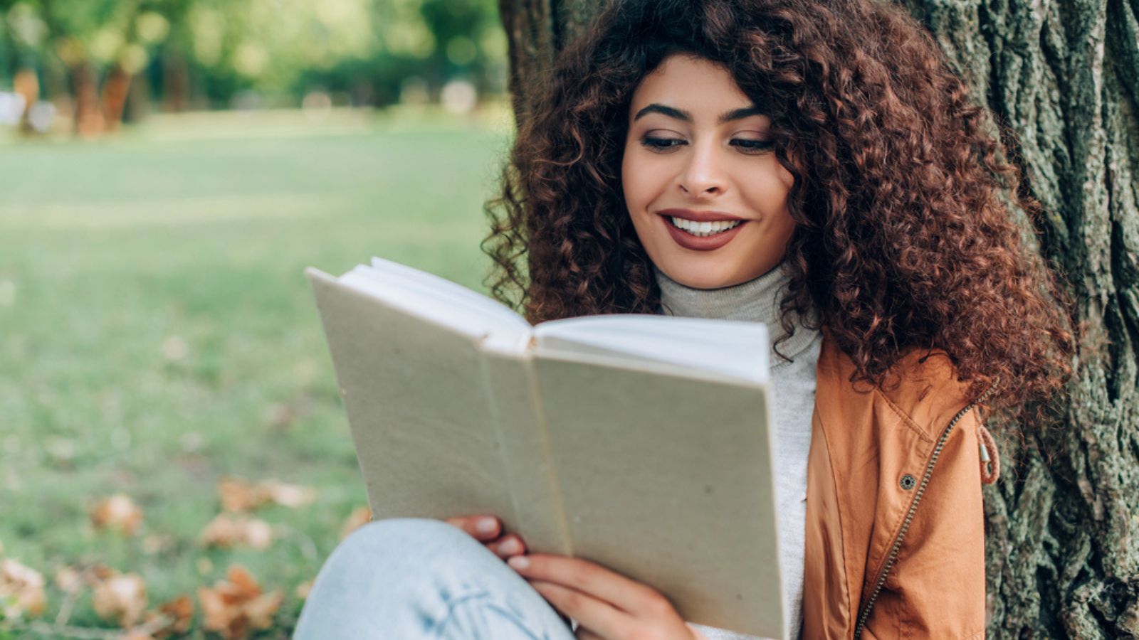 Selective focus of curly woman in autumn outfit reading book in park