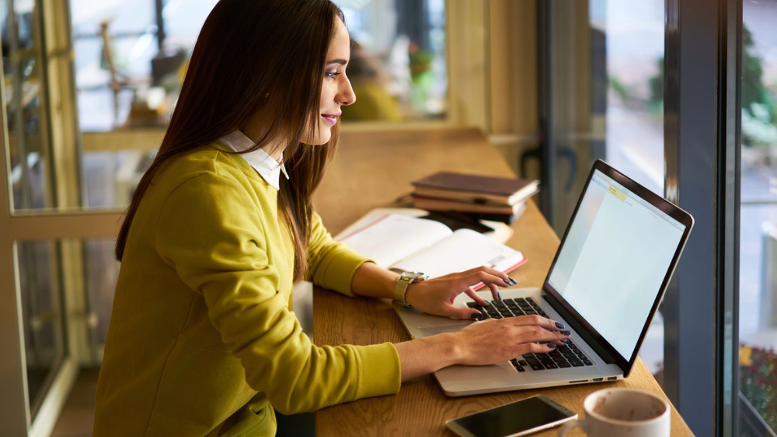 Positive young female journalist sitting with pen in hand at table with markers notebook and working on laptop while looking at screen during online information gathering for publication in cafe