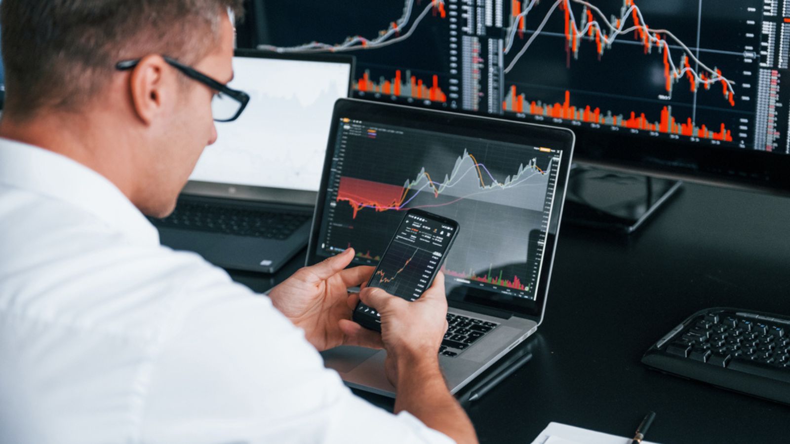 Young man stock broker works indoors in the office. Many of displays laptop and using phone
