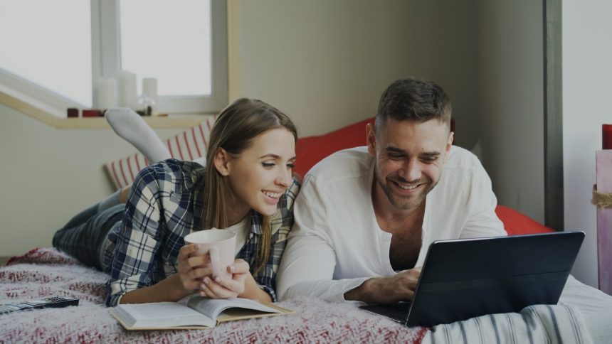 Couple lying on bed with laptop and coffee; budgeting as a couple