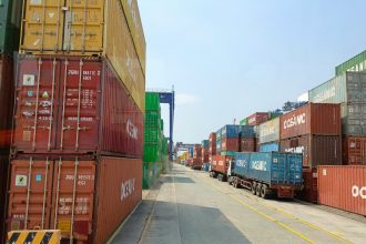 a long line of shipping containers on the side of a road; tariffs